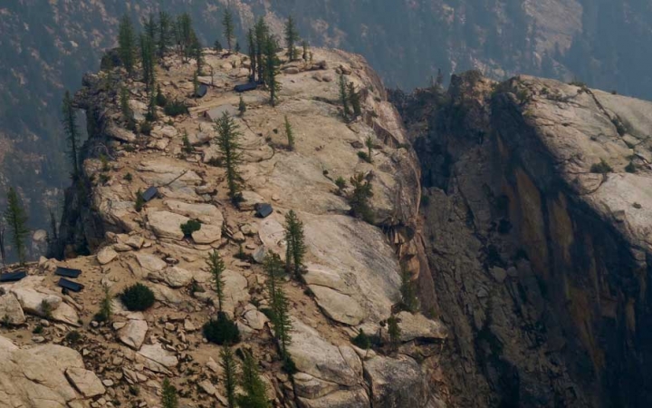 From an aerial point of view, a couple of tents rest on a cliff high above the landscape below. A few evergreen trees also dot the cliff. 